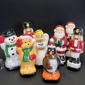 Puede incluir: Una colección de figuras navideñas de cerámica, que incluyen un muñeco de nieve, un oso, un ángel, un pingüino, Papá Noel y cascanueces. Las figuras son de colores brillantes con detalles festivos, como bufandas y sombreros. El fondo es negro.