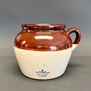 May include: A vintage stoneware crock with a brown glazed top and a cream-colored base. It features a rounded body, a handle, and a blue emblem with the number "3" on the front. The crock is likely used for food storage.