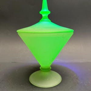 Op de afbeelding: Een felgroene glazen bonbonnière met een conische body en een bijpassende deksel. De schaal staat op een ronde voet en heeft een puntige finial op het deksel. Het glas heeft een licht matte uitstraling.