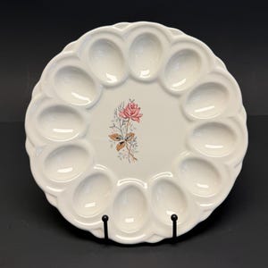 May include: White ceramic devilled egg serving platter with scalloped edges and 12 egg-shaped indentations. A pink rose and floral design is in the centre. The platter is displayed on a black metal stand against a black background.