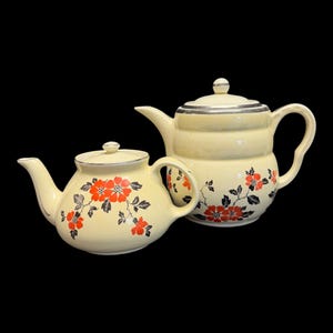 Op de afbeelding: Twee crèmekleurige vintage theepotten met bloemmotieven. De theepotten hebben rode bloemen en zwarte bladeren. Eén theepot heeft een zilveren rand en deksel. De theepotten staan tegen een zwarte achtergrond.