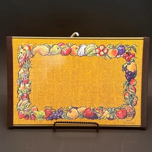 May include: A rectangular, vintage-style decorative board with a yellow burlap-textured background. The board is framed with a brown border and adorned with a colorful border of various fruits. A white hook is attached to the top.