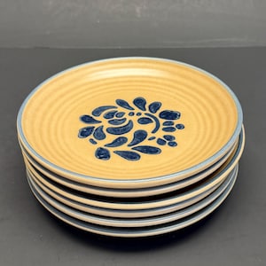 May include: A stack of six ceramic plates. Each plate has a tan center with a blue floral design and a blue rim. The plates have a ridged texture.