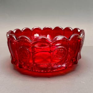 Puede incluir: Un cuenco de vidrio rojo vibrante con un borde festoneado. El cuenco tiene forma circular con diseños en relieve en los lados. El vidrio es de un rojo profundo y transparente. El cuenco se asienta sobre una base plana.