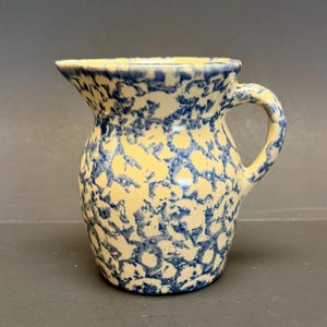 Puede incluir: Una jarra de cerámica pequeña con una base color crema y un patrón azul moteado. La jarra tiene un cuerpo redondeado, un asa curva y un pico vertedor. El diseño recuerda a la cerámica antigua.