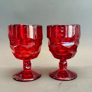 Könnte beinhalten: Zwei rubinrote Vintage-Becher. Die Gläser haben ein facettiertes Design auf der Schale und einen Stiel mit rundem Fuß. Die Becher sind aus Glas und werden auf einer grauen Oberfläche präsentiert.