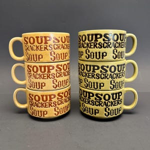 May include: Six vintage soup mugs stacked in two sets of three. The mugs are beige with brown and black lettering that reads "Soup Crackers Soup". Each mug has a handle.