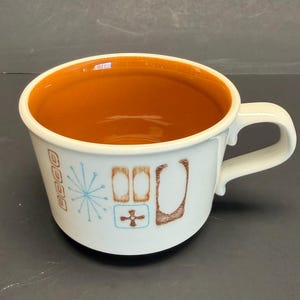 Puede incluir: Taza de cerámica blanca con asa, con interior naranja quemado y un patrón decorativo en marrón y azul. La taza tiene un diseño moderno de mediados de siglo.