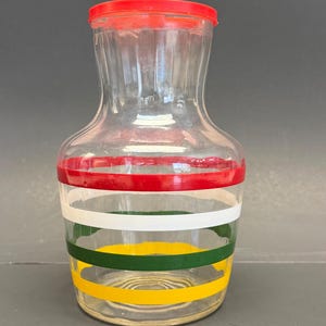 Op de afbeelding: Een heldere glazen kan met een rode deksel en kleurrijke horizontale strepen. De strepen zijn rood, wit, groen en geel. De kan heeft een brede basis en een smallere hals.