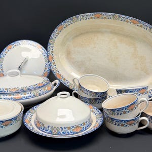 May include: A ceramic dinnerware set featuring an off-white base with a decorative border of blue scrolls and orange floral accents. The collection includes a large oval platter, a lidded tureen, a lidded serving dish, and several matching tea cups.