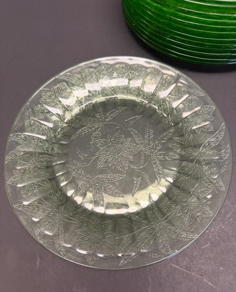 Puede incluir: Un plato de vidrio transparente con un patr&oacute;n floral en relieve de flores y hojas de pascua en toda la superficie. El plato tiene un borde festoneado y un borde ligeramente elevado. Se ve una parte de un objeto de vidrio verde oscuro.