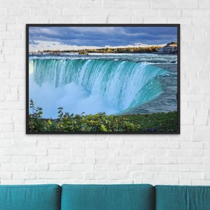 Puede incluir: Una impresión enmarcada de las Cataratas del Niágara, que muestra el agua en cascada en tonos azules y turquesas. El cielo es una mezcla de azul y gris, con toques de follaje otoñal. La obra de arte está sobre un sofá turquesa.