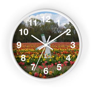 Könnte beinhalten: Weiße Wanduhr mit einem Landschaftsbild einer Windmühle in einem Feld mit bunten Tulpen. Das Zifferblatt hat schwarze Ziffern und Zeiger mit einem weißen Rahmen. Der Hintergrund zeigt einen blauen Himmel und Bäume.