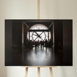May include: A framed print of a large clock inside a building. The clock is round with Roman numerals and is backlit. Silhouetted figures of people are visible in the foreground, and the print is displayed on an easel.