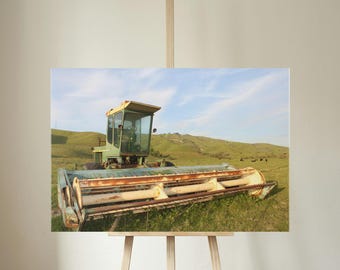 May include: A photograph of a vintage green and yellow combine harvester in a field. The machine has a large cutting head and a cab with a person inside. The background shows rolling green hills and a blue sky.