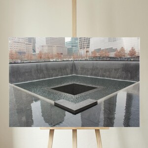 May include: A photograph of the 9/11 Memorial in New York City. The image shows the reflecting pools with the names of the victims inscribed around the edges. The surrounding buildings are reflected in the water.