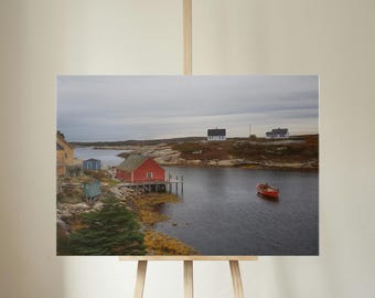 May include: A landscape print featuring a coastal scene with a red building on stilts, a small red boat, and several other buildings. The sky is overcast, and the water is calm. The print is displayed on a wooden easel.