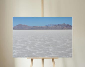 May include: A landscape print featuring a vast, white salt flat under a clear blue sky. Distant mountains in shades of brown and purple provide a backdrop. The print is displayed on a wooden easel.