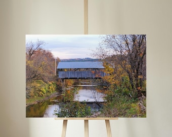 Op de afbeelding: Een landschapsfoto van een overdekte brug over een rustige rivier. De brug is gemaakt van verweerd hout met een metalen dak. De omringende bomen tonen herfstkleuren, met gele en oranje bladeren.