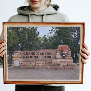 以下が含まれることがあります： グランドキャニオン国立公園の入り口の看板の写真が額縁に入っています。石でできた看板には公園の名前と国立公園局のエンブレムが描かれています。画像は木々と曇り空を背景にしています。