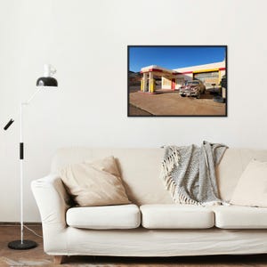 May include: A framed photograph of a vintage gas station with a classic car parked in front. The gas station has a red and yellow awning, and the sky is clear blue. The car is a light brown color.