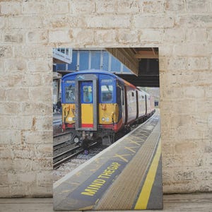 Puede incluir: Impresión en lienzo de un tren en una estación. El tren es azul, amarillo y rojo. El andén tiene líneas amarillas y el texto "MIND THE GAP". La impresión está apoyada contra una pared de ladrillos.