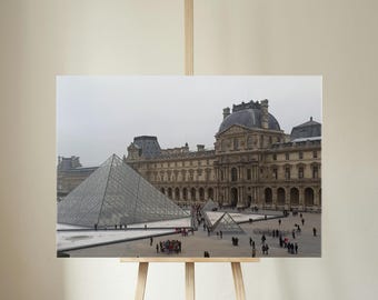 May include: A framed print of the Louvre Museum in Paris, France, featuring the iconic glass pyramid and the historic palace. The image is set against a cloudy sky, with people visible in the courtyard.