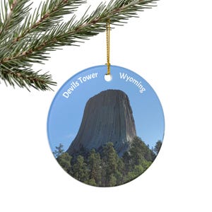 Op de afbeelding: Een ronde kerstversiering met Devils Tower in Wyoming. De versiering heeft een blauwe achtergrond met de tekst "Devils Tower Wyoming". De afbeelding toont de toren boven een bos.
