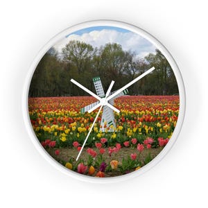 Könnte beinhalten: Weiße Wanduhr mit einem Bild einer Windmühle in einem Feld mit bunten Tulpen. Das Zifferblatt zeigt eine Landschaft mit roten, gelben und orangen Tulpen. Die Uhr hat ein weißes Gehäuse und schwarze Zeiger, ideal für Wohnzimmer oder Küche.