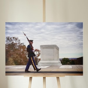May include: A framed print of a soldier in uniform carrying a rifle, walking past a memorial. The soldier is wearing a dark blue coat with gold accents and blue pants with a gold stripe. A wreath is placed near the memorial.