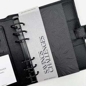 Könnte beinhalten: Schwarzer Lederplaner mit silbernen Akzenten. Der Planer ist geöffnet und zeigt eine silberne Karte mit dem Text "SAVINGS CHALLENGES" und einen schwarzen Einsatz mit Blättermuster. Für Budget-Essentials.