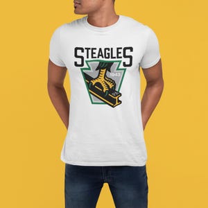 Könnte beinhalten: Weißes T-Shirt mit dem Text "STEAGLES" über einem Logo mit einem grünen und schwarzen Adlerklauen-Design, dem Jahr "1943" und den Buchstaben "PA". Das T-Shirt hat kurze Ärmel.