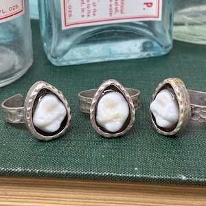 Puede incluir: Tres anillos de plata, cada uno con una muela blanca engastada en un bisel en forma de lágrima. Los anillos descansan sobre un libro verde, con botellas de veneno vintage al fondo.