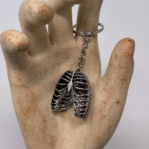 May include: A silver-colored keychain featuring a detailed rib cage design. The keychain is attached to a silver ring and chain. The rib cage pendant is made of metal and has a three-dimensional appearance. The background is a neutral color.
