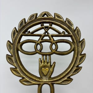 May include: A bronze-colored, decorative metal object with a circular design. The design includes a hand with a heart, a series of connected circles, and a laurel wreath. The object has a handle at the bottom.
