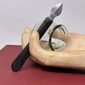 May include: A small, silver-bladed knife with a black handle rests on a clear glass object, held by a beige, hand-shaped sculpture. The sculpture sits on a red surface. The knife is angled towards the viewer.