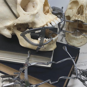 May include: Two human skulls with dental instruments attached, resting on a stack of dark books and anatomical diagrams. The skulls are positioned at different angles, showcasing the dental tools. The image has a vintage, medical aesthetic.