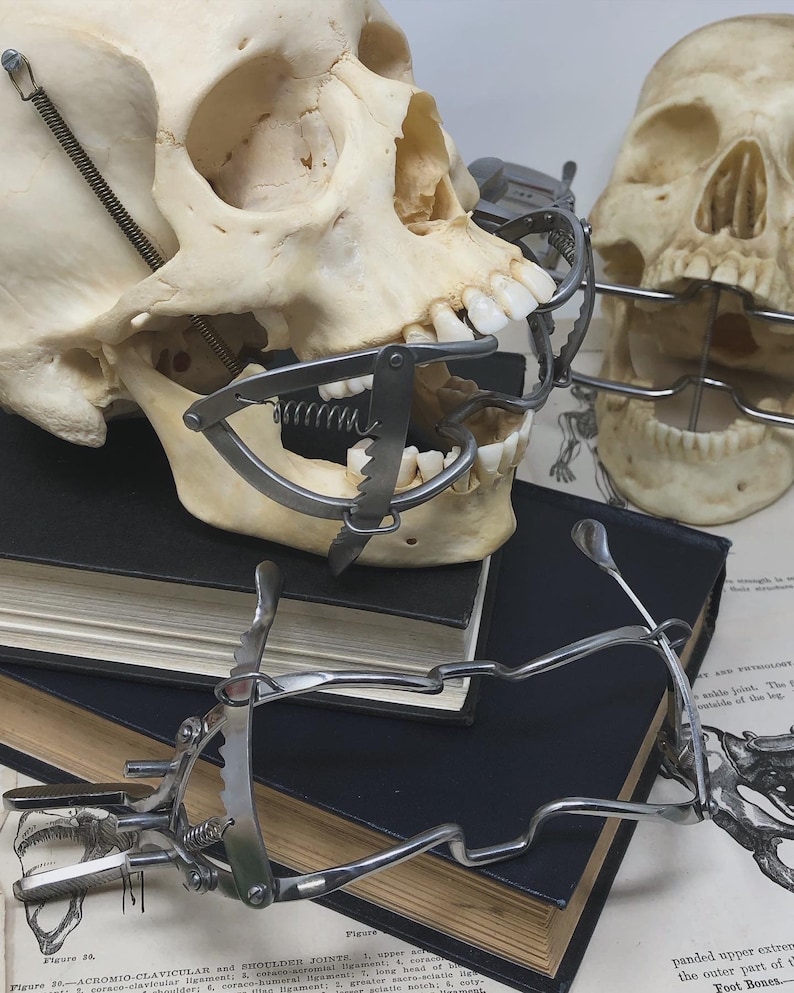 May include: Two human skulls with dental instruments attached, resting on a stack of dark books and anatomical diagrams. The skulls are positioned at different angles, showcasing the dental tools. The image has a vintage, medical aesthetic.