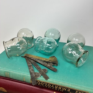 May include: A selection of clear and teal glass cupping therapy cups with a rounded form and flared openings. Several metal tools are arranged on a stack of books with teal and burgundy covers. The background is plain white.