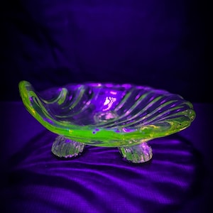 May include: A clear, three-footed glass dish with a shell-like design. The dish has a vibrant green rim and accents, with a swirling pattern throughout. The dish is set against a dark purple background.