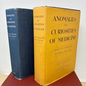 Op de afbeelding: Twee vintage boeken getiteld "Anomalies and Curiosities of Medicine" door George M. Gould en Walter L. Pyle. Eén boek is marineblauw, de andere is geel. De boeken liggen op een rode ondergrond.