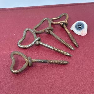 May include: Four antique metal screw eyes with heart-shaped handles and a prosthetic eye on a red surface. The screw eyes are made of aged metal with a threaded shaft. The prosthetic eye is white, blue, and pink.