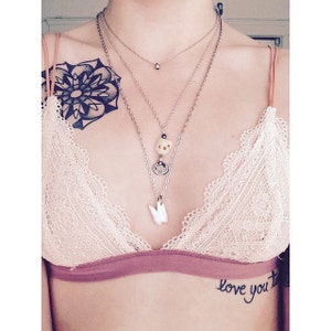 May include: A woman wearing a lacy bra with a black mandala tattoo on her left shoulder. She is wearing multiple necklaces with charms, including a tooth charm. The text "love you to" is tattooed on her right side.
