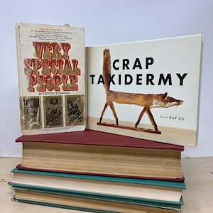 Puede incluir: Una pila de libros antiguos con cubiertas rojas, turquesas y verdes. El libro superior está abierto, mostrando una ilustración de un zorro disecado y el título "CRAP TAXIDERMY". Otro libro se titula "VERY SPECIAL PEOPLE".
