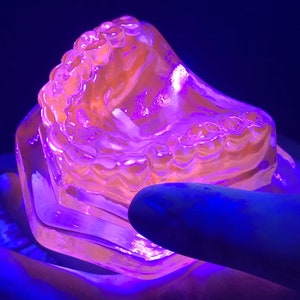 May include: A clear plastic dental mold with a purple glow under a blacklight. The mold is shaped like a set of teeth and is being held in a hand.