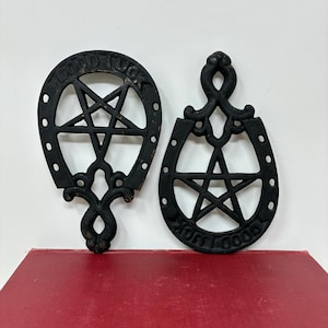May include: Two black cast iron trivets featuring a horseshoe shape with a central five-pointed star design. Each piece has the words GOOD LUCK embossed along the curved edge. The metal surfaces show a dark, matte finish with visible texture.