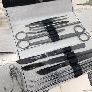 Puede incluir: Una caja de cuero negro con un forro de lona blanca, que contiene un conjunto de herramientas quirúrgicas. Las herramientas incluyen escalpelos, pinzas, tijeras y ganchos. La caja está abierta y las herramientas son visibles.