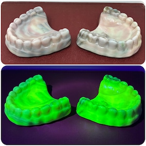 May include: Two sets of false teeth, one pair in shades of white and gray, the other pair glows bright green. The teeth are arranged in a curved shape, with individual tooth details visible. The background is a dark purple.