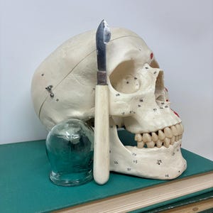 May include: A medical display featuring a human skull model, a small scalpel with a bone handle, and a glass cupping jar. The skull rests on a stack of green and brown books, with a white background.