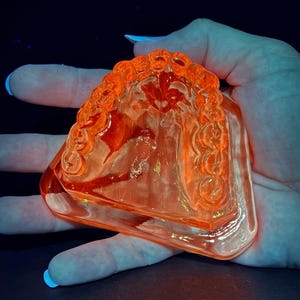 May include: A glowing orange glass object with a decorative, curved top and a rectangular base. The glass has a textured surface and is held in a hand against a dark background. The object appears to be a decorative piece.